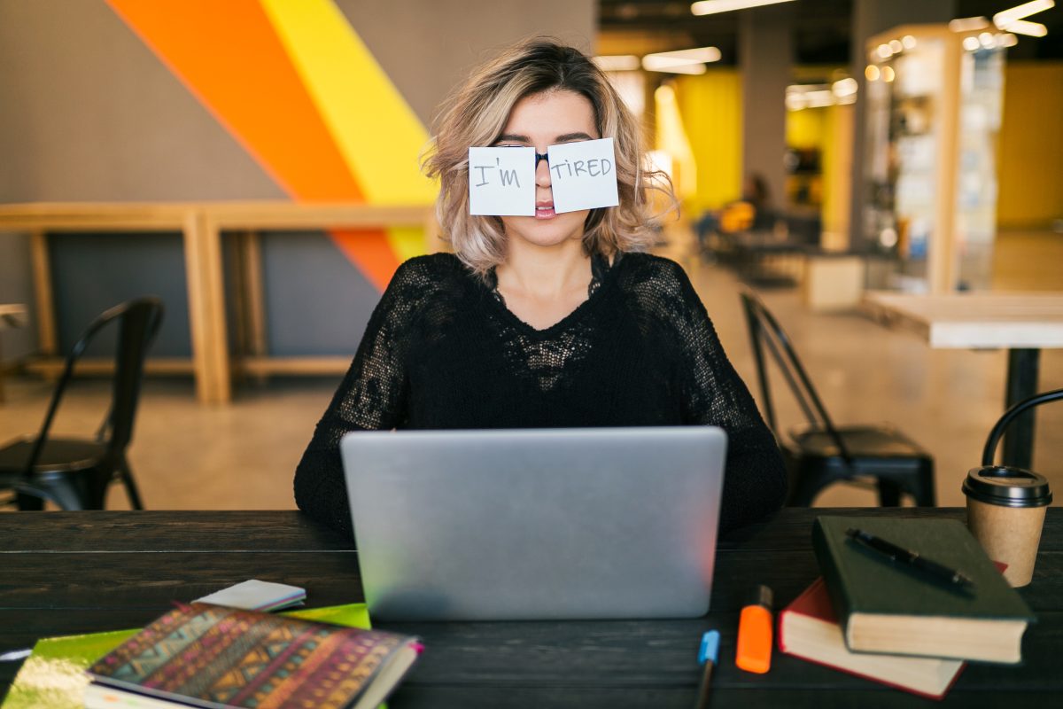 portrait of young pretty tired woman with paper stickers on glasses sitting at table in black shirt working on laptop in co-working office, funny face expression, frustrated emotion