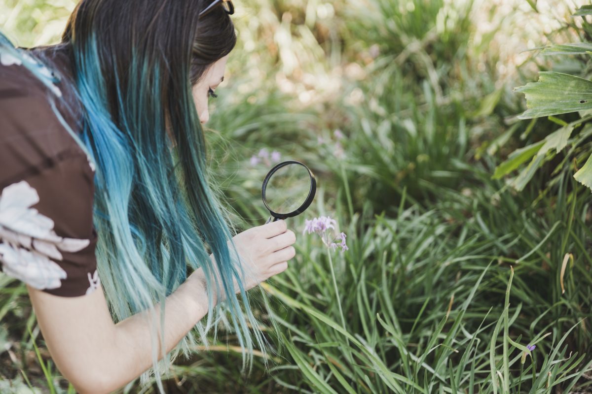 Woman with cool blue hair looking at a flower through a magnifying glass.